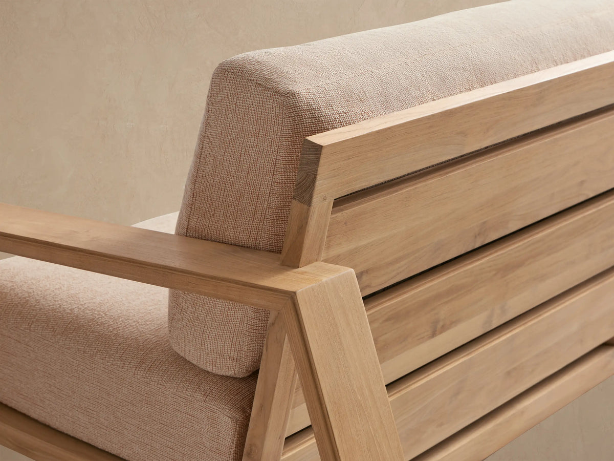 Canyon Outdoor Teak Sofa, natural wood frame, tan cushions, on a paved patio against a light wall with shadows.