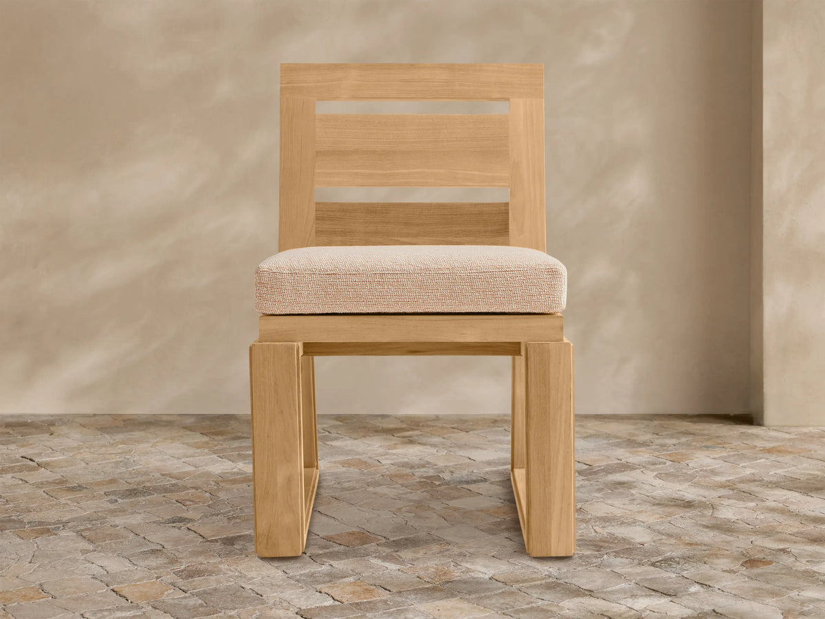 Canyon Outdoor Teak Dining Side Chair with natural wood frame, slatted back, and light beige cushion, angled on a stone floor.