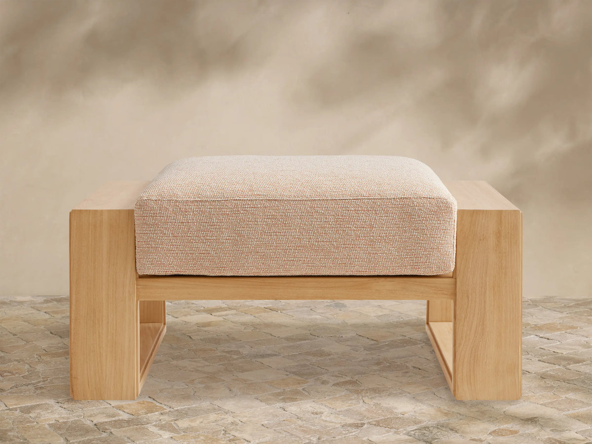 Canyon Outdoor Teak Ottoman, front view. Natural teak wood frame with a rectangular, textured beige cushion. On a stone patio.
