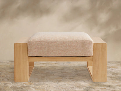 Canyon Outdoor Teak Ottoman, front view. Natural teak wood frame with a rectangular, textured beige cushion. On a stone patio.