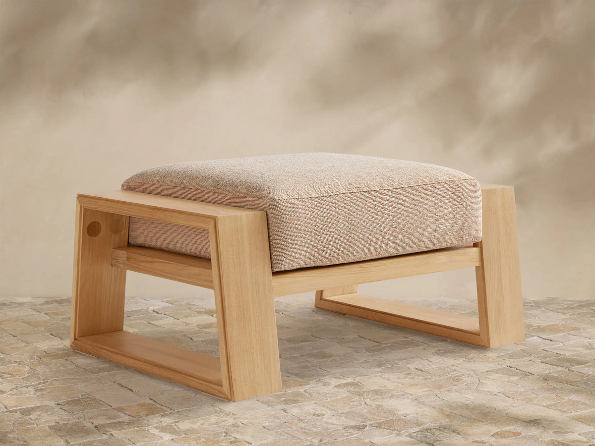 Canyon Outdoor Teak Ottoman, front view. Natural teak wood frame with a rectangular, textured beige cushion. On a stone patio.