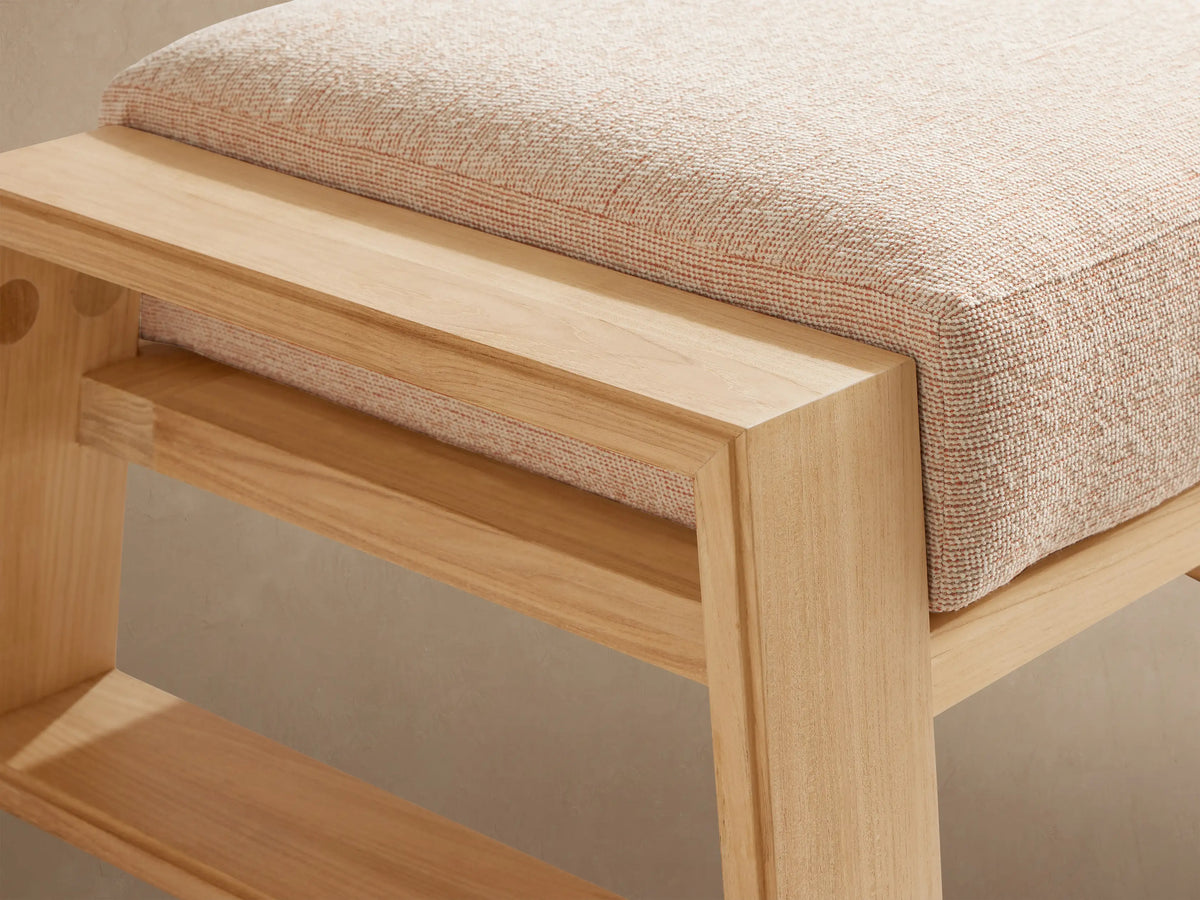 Canyon Outdoor Teak Ottoman with a light beige cushioned top on a natural light brown teak wood frame, on a stone patio.