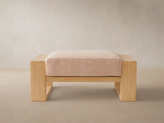 Canyon Outdoor Teak Ottoman, front view. Natural teak wood frame with a rectangular, textured beige cushion. On a stone patio.