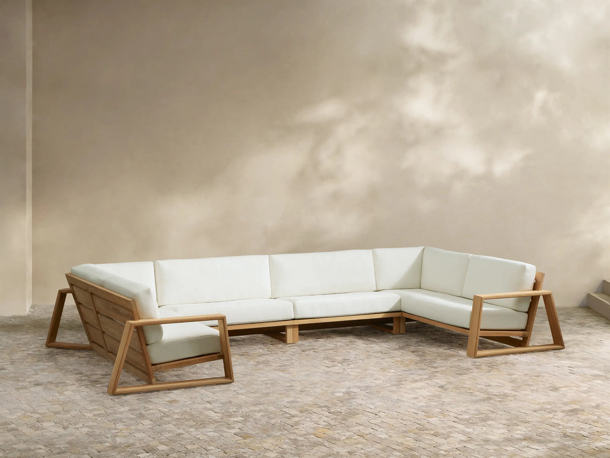 U-shaped Canyon Outdoor Teak Four-Piece Sectional with natural teak frame & white cushions on a patio, wall behind with cloud shadows.