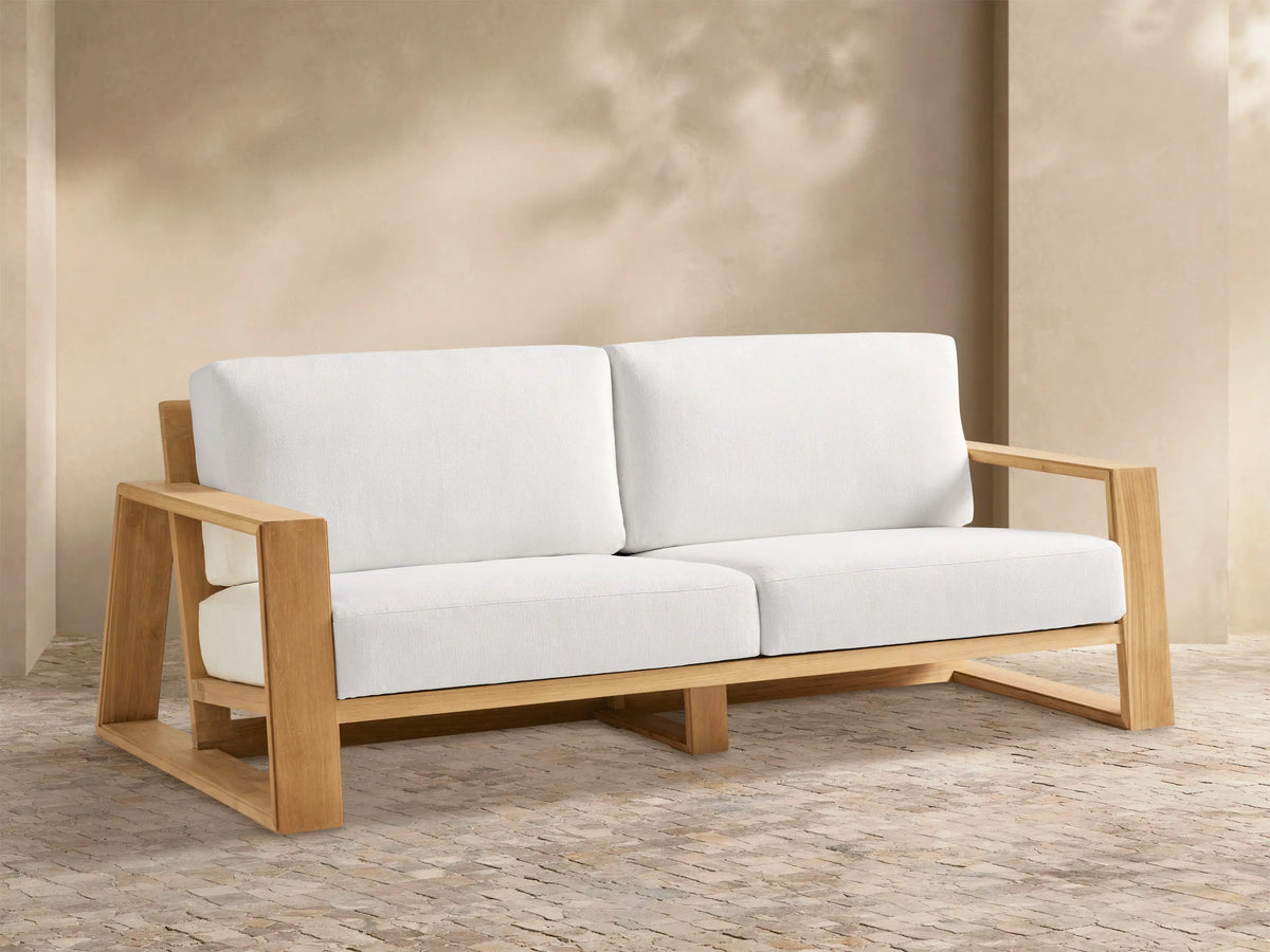Canyon Outdoor Teak Sofa: 2-seater with a natural teak wood frame and white cushions, set on a textured patio.