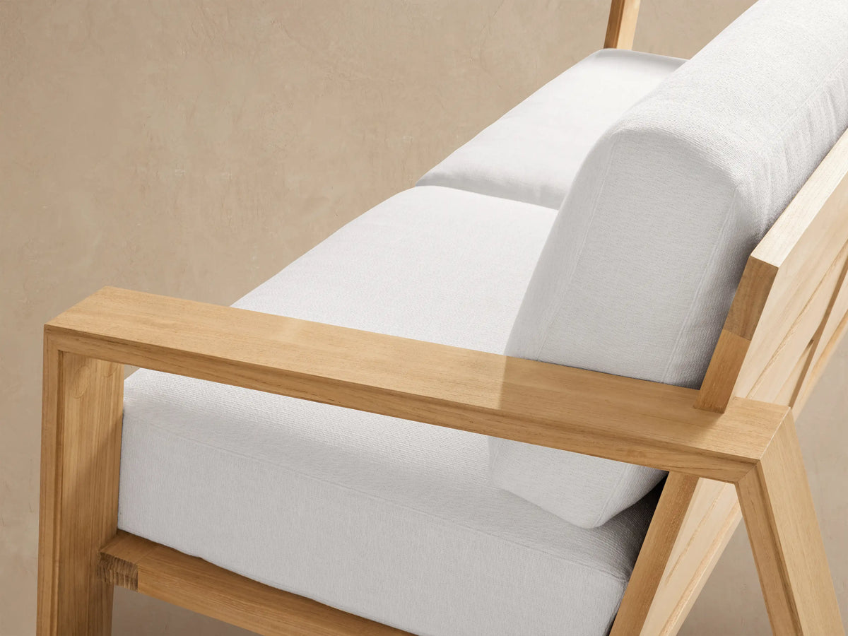 Canyon Outdoor Teak Sofa, angled right, with a natural teak wood frame and plush white cushions on a paved patio.