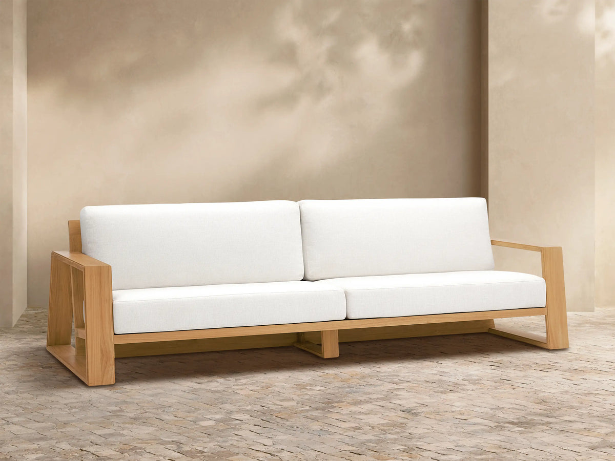 Canyon Outdoor Teak Sofa with a light natural wood frame and two white seat and back cushions, on a stone patio.