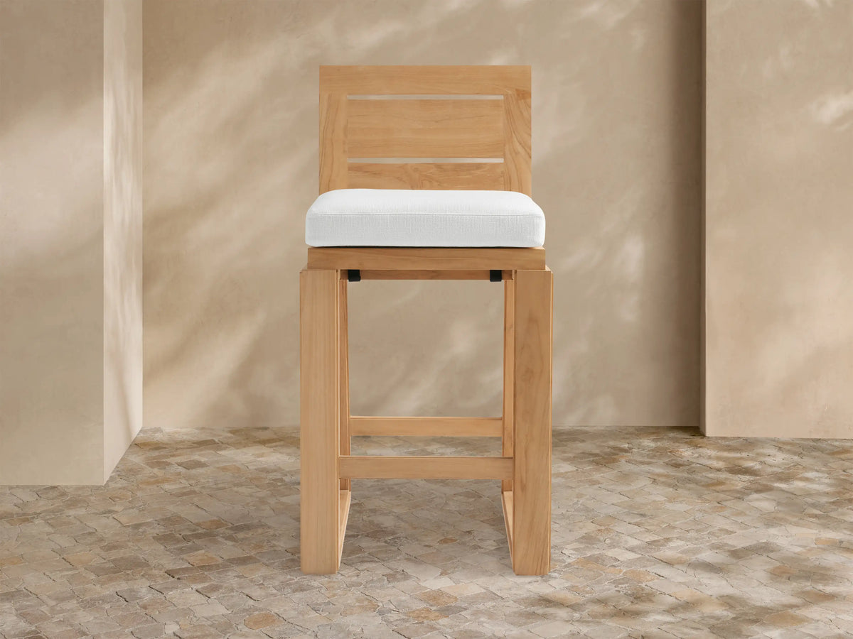 Canyon Outdoor Teak Barstool with slatted back and white cushion, standing on a mosaic patio with dappled sunlight on the wall.