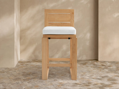 Canyon Outdoor Teak Barstool with slatted back and white cushion, standing on a mosaic patio with dappled sunlight on the wall.