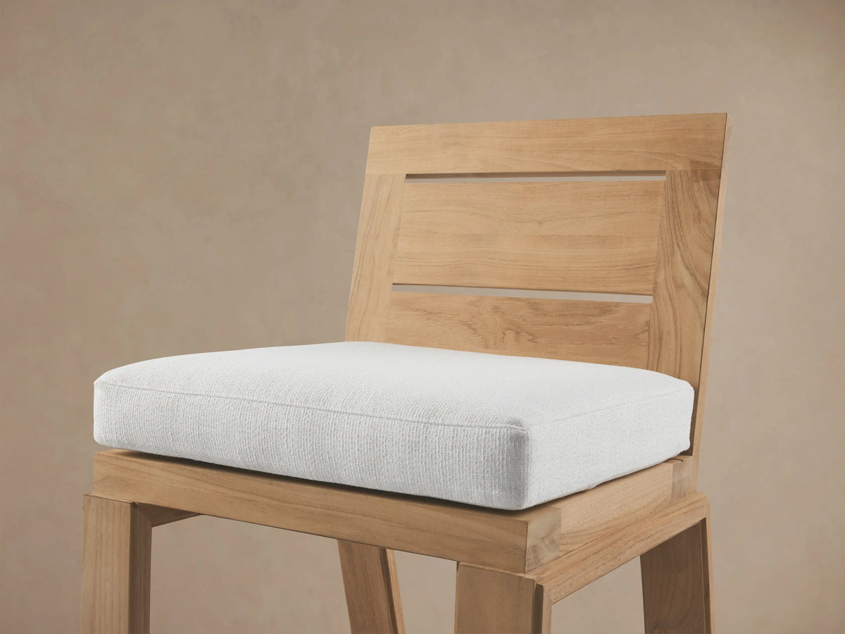Canyon Outdoor Teak Barstool with light wood finish, white seat cushion, slatted back, on stone patio with dappled light.
