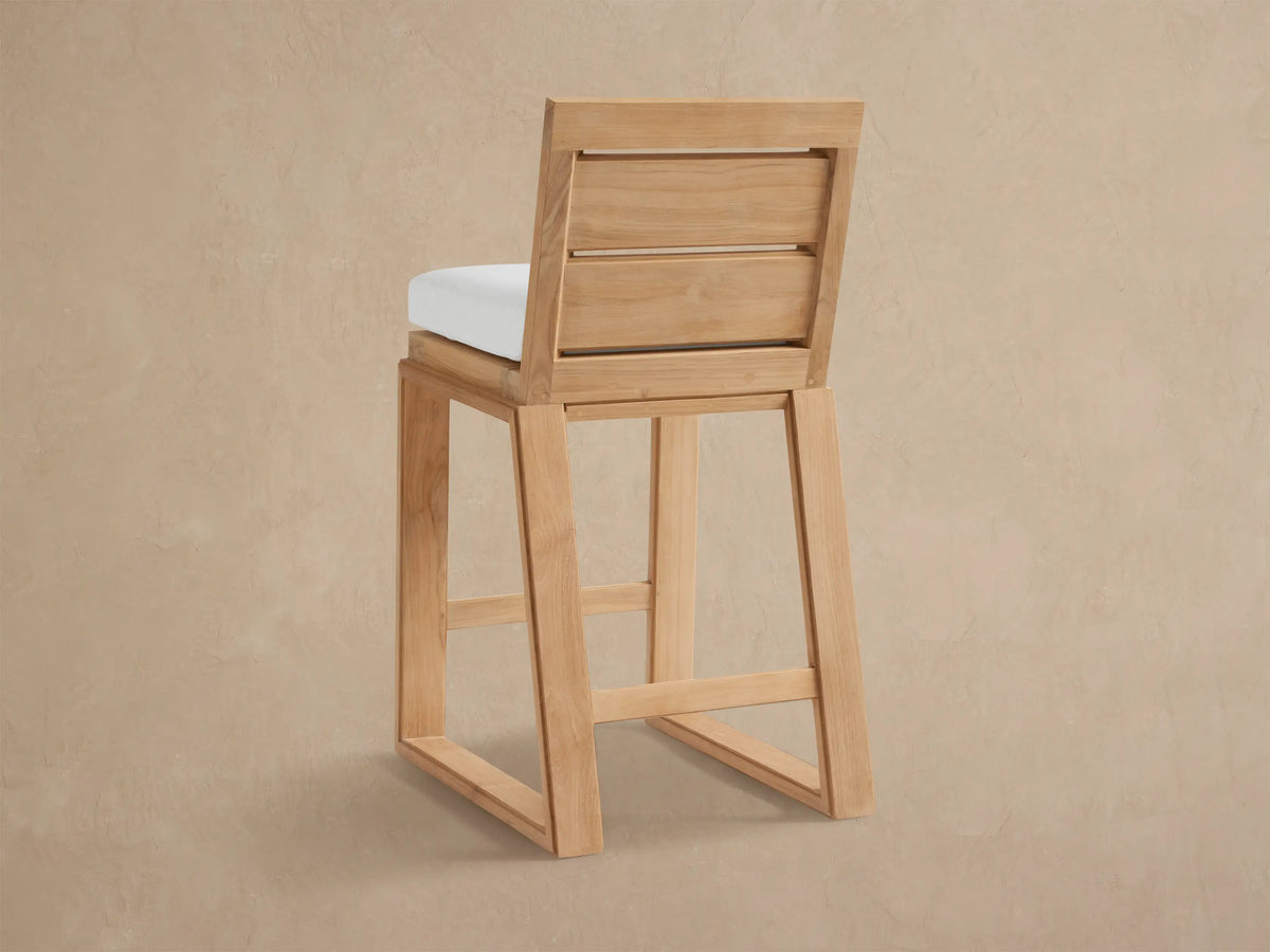 Canyon Outdoor Teak Barstool. Angled view of light natural teak wood frame with slatted back and thick, textured off-white cushion.