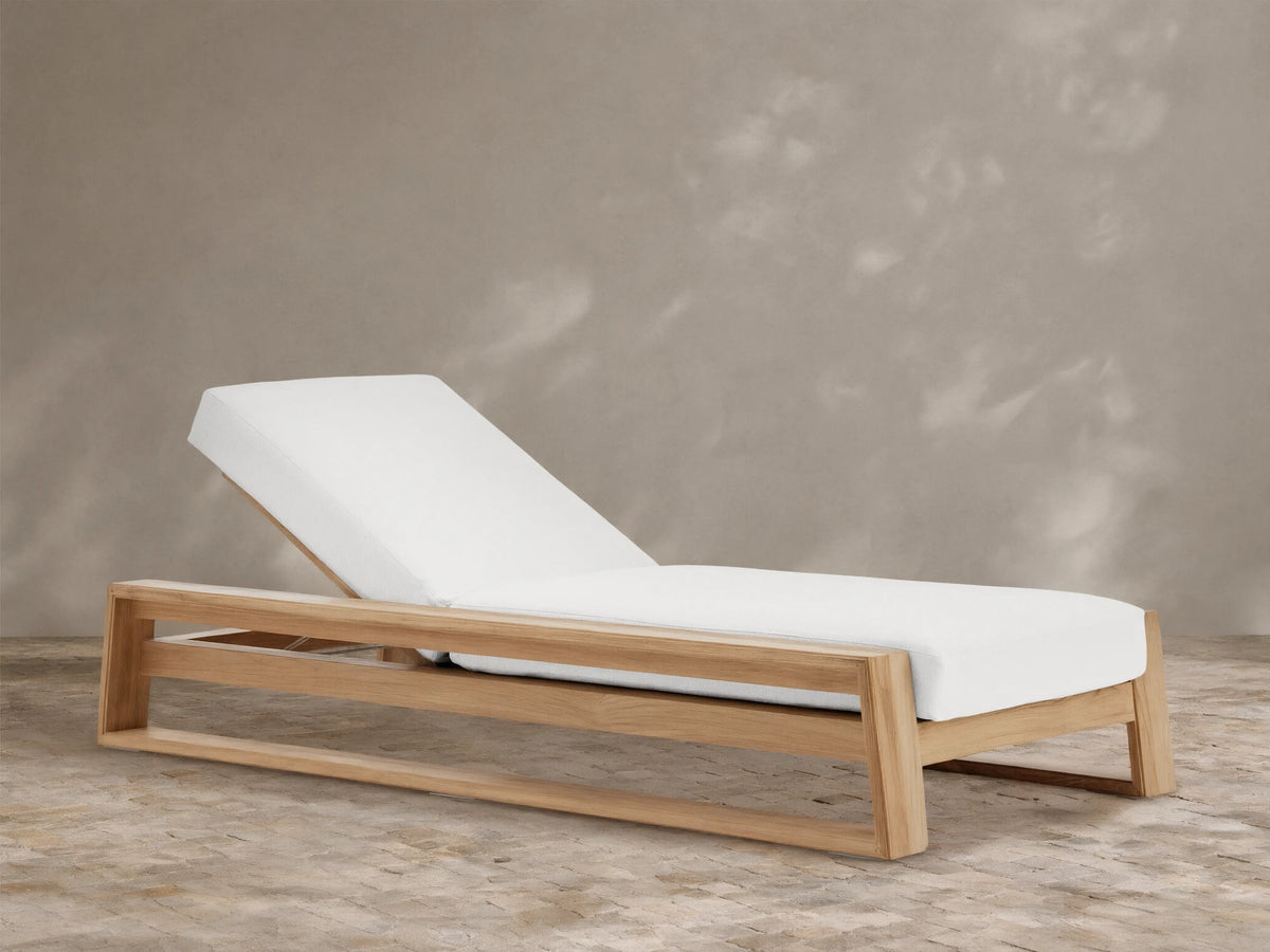 Canyon Outdoor Teak Chaise with natural wood frame and plush white cushion, back reclined, on a stone patio against a tan wall.