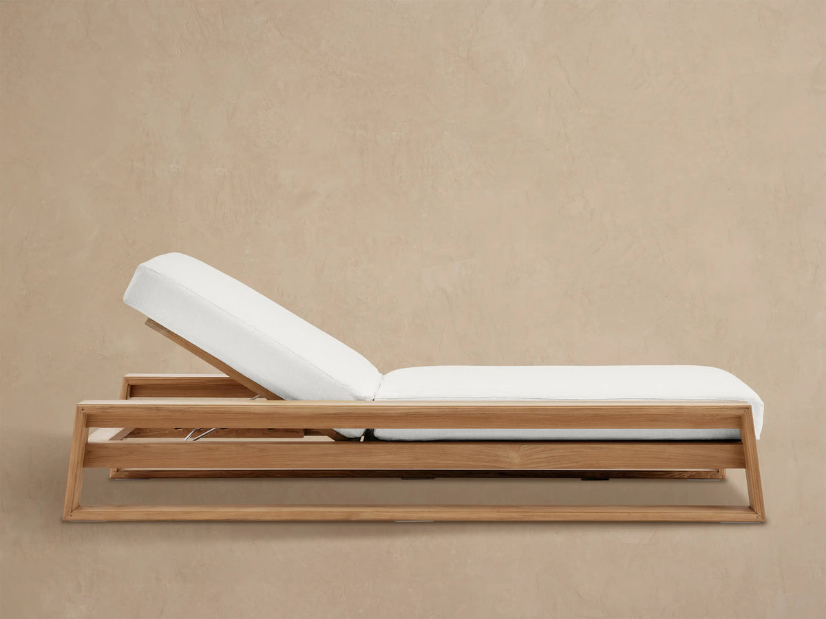 Canyon Outdoor Teak Chaise with natural wood frame and plush white cushion, back reclined, on a stone patio against a tan wall.