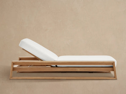 Canyon Outdoor Teak Chaise with natural wood frame and plush white cushion, back reclined, on a stone patio against a tan wall.