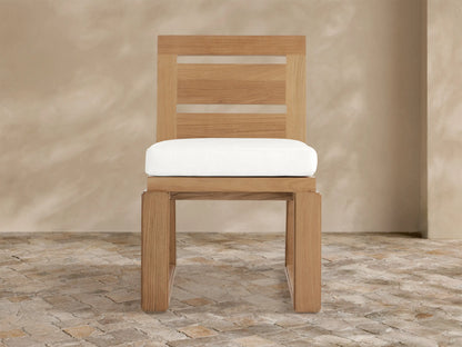 Canyon Outdoor Teak Dining Side Chair. Light brown teak wood with slatted back, white cushion, on stone patio against textured wall.