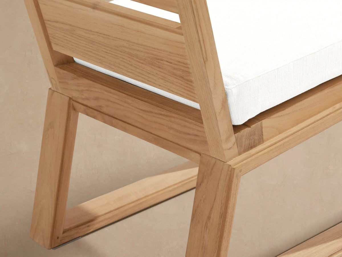 Canyon Outdoor Teak Dining Side Chair, front view. Natural teak frame, slatted back, white cushion. Beige wall, stone floor.