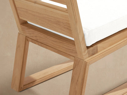 Canyon Outdoor Teak Dining Side Chair, front view. Natural teak frame, slatted back, white cushion. Beige wall, stone floor.
