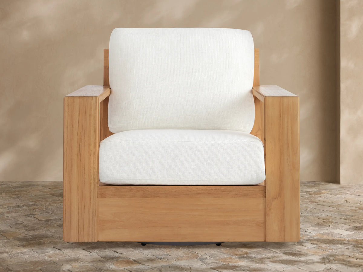 Canyon Outdoor Teak Swivel Chair, natural light teak wood frame & thick white cushions. Angled view on a textured stone patio.