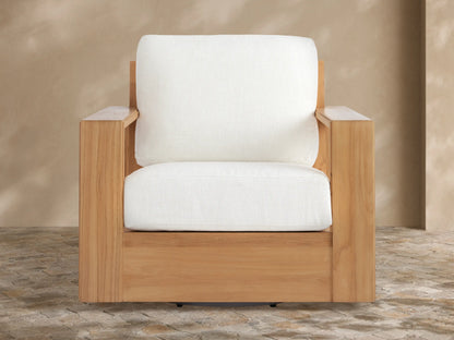 Canyon Outdoor Teak Swivel Chair, natural light teak wood frame & thick white cushions. Angled view on a textured stone patio.
