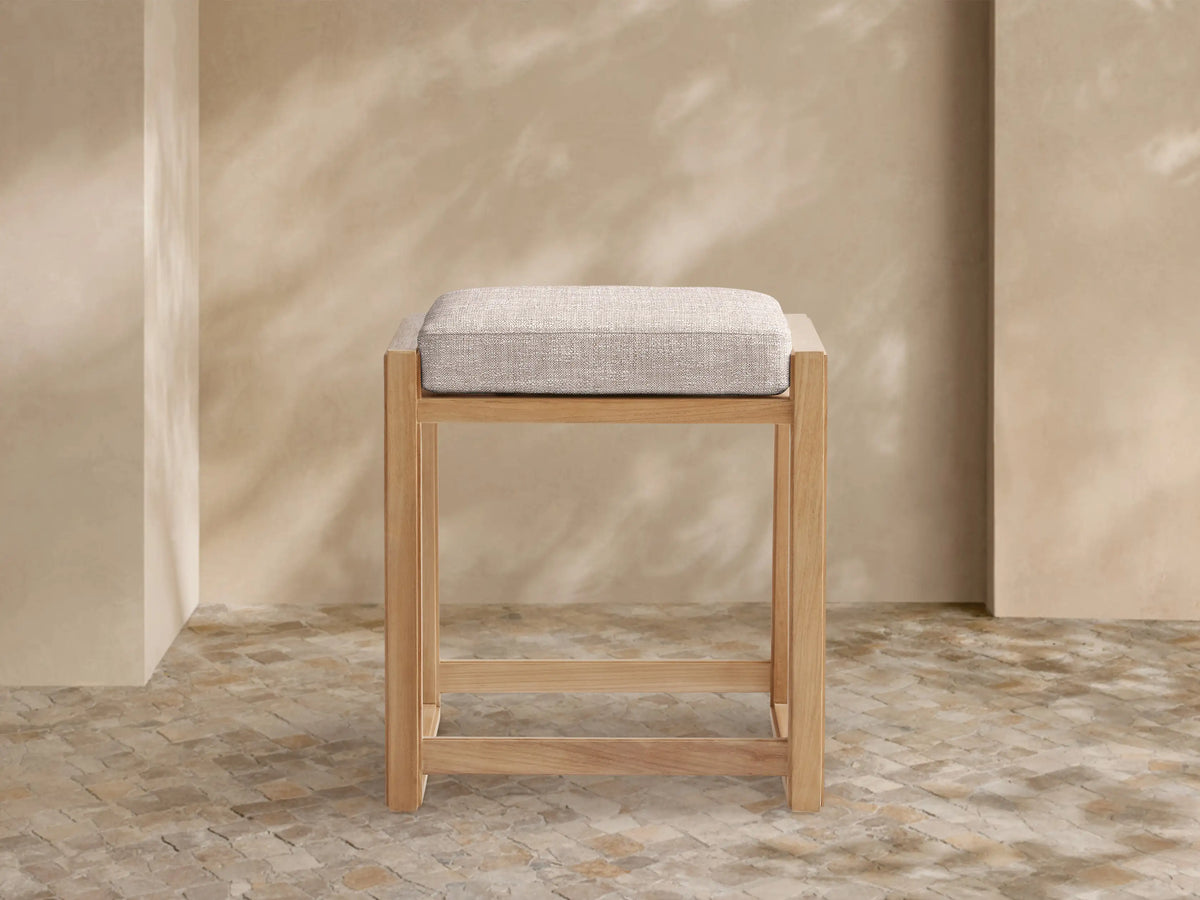 Canyon Outdoor Teak Backless Counter Stool, light natural teak wood frame, rectangular, with a light beige fabric cushion, on tiled floor.