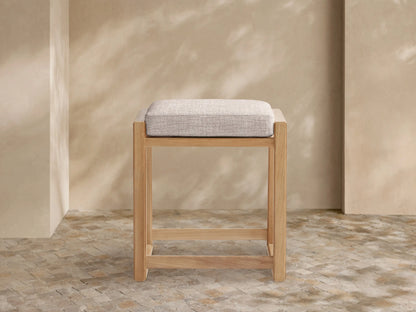Canyon Outdoor Teak Backless Counter Stool, light natural teak wood frame, rectangular, with a light beige fabric cushion, on tiled floor.