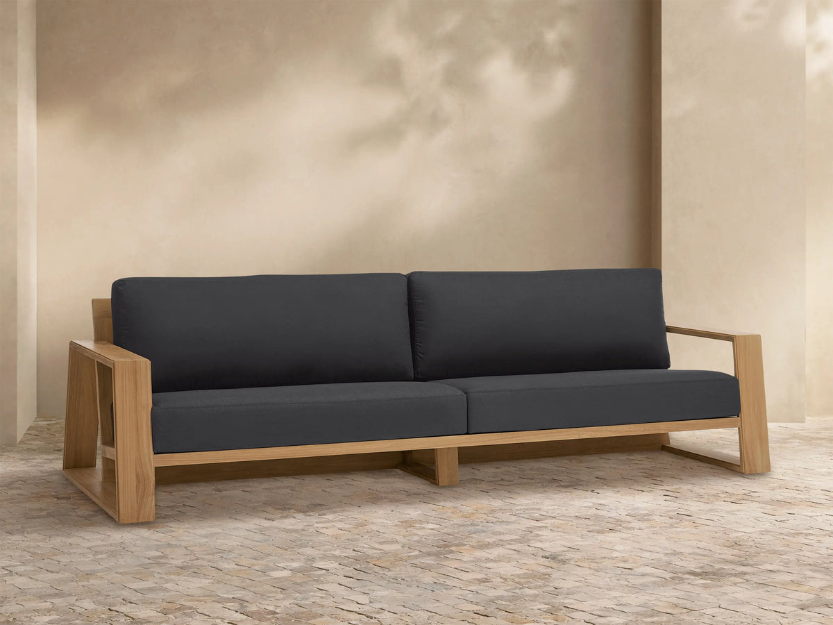 Canyon Outdoor Teak Sofa with natural wood frame and dark gray cushions, set against a light tan wall on a tiled patio.