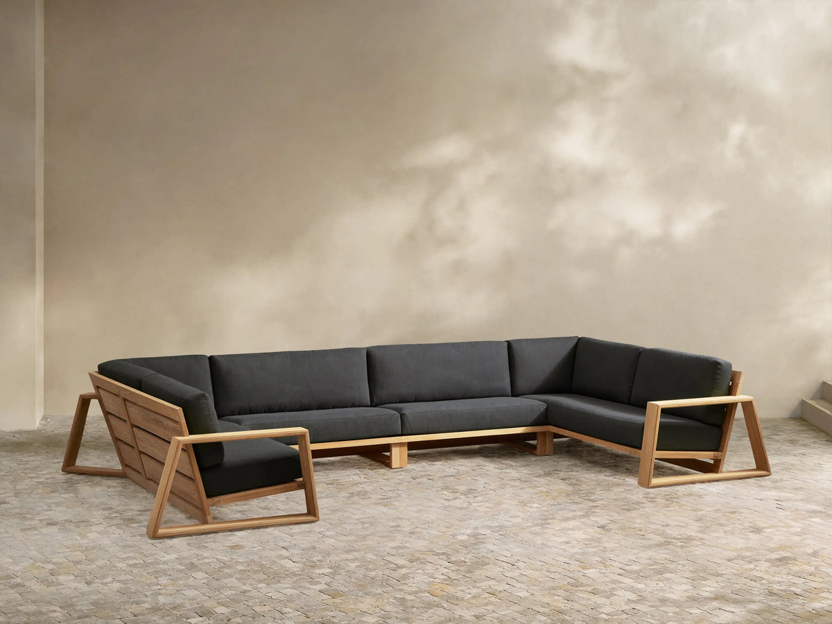 U-shaped Canyon Outdoor Teak Four-Piece Sectional with black cushions, natural teak frame, on stone pavers against a light wall.