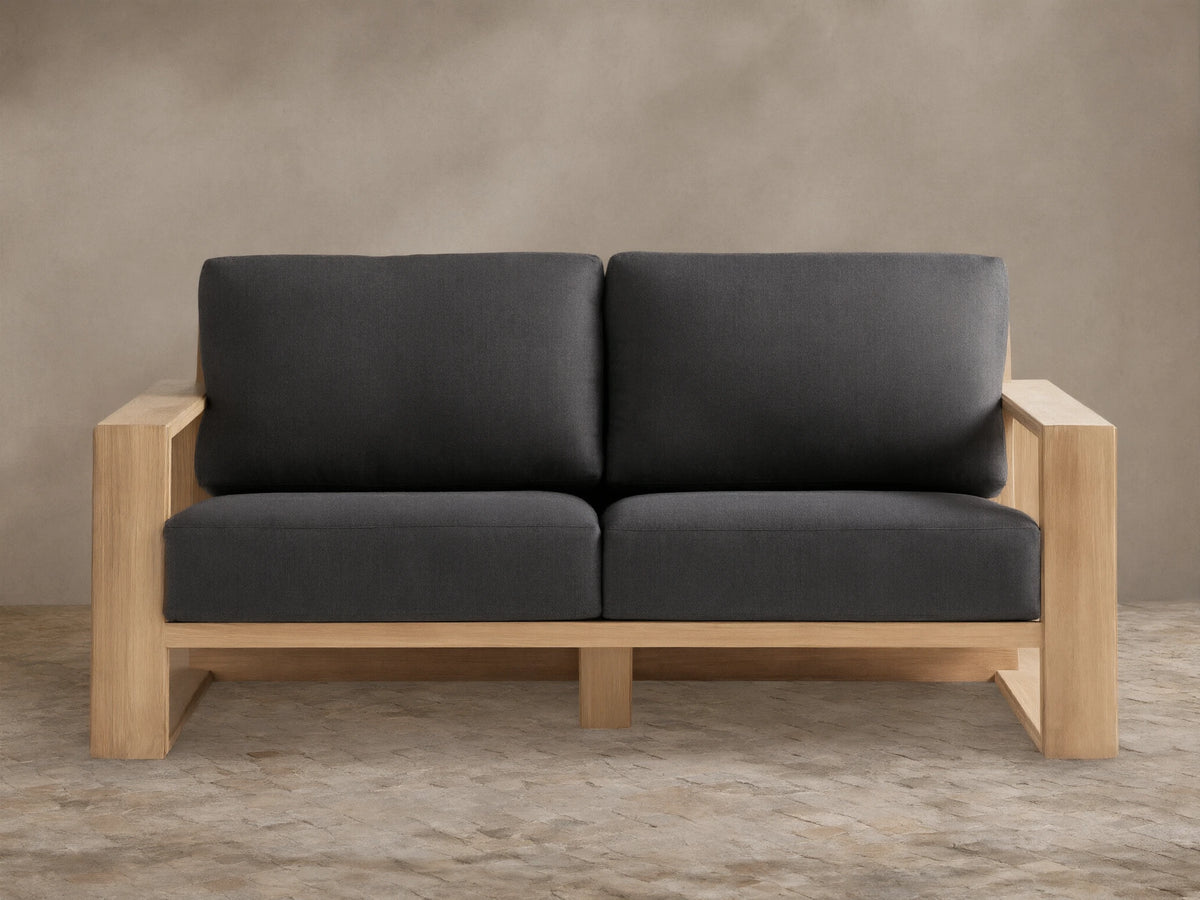 Canyon Outdoor Teak Sofa with natural teak wood frame and two dark gray cushions on a stone patio against a tan wall.