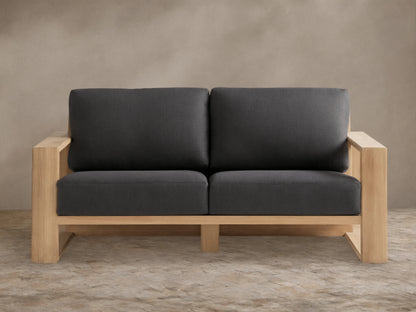 Canyon Outdoor Teak Sofa with natural teak wood frame and two dark gray cushions on a stone patio against a tan wall.