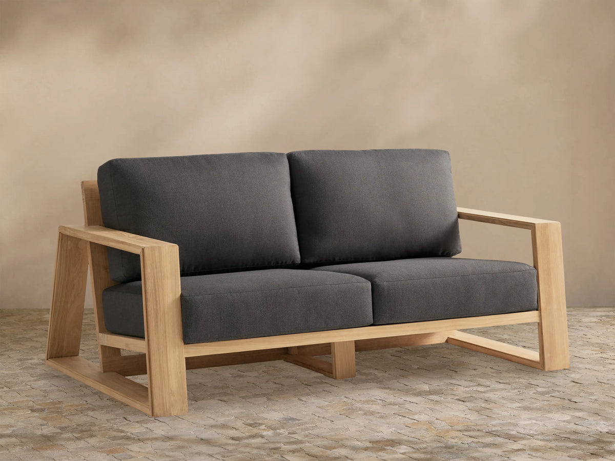 Canyon Outdoor Teak Sofa with natural teak wood frame and two dark gray cushions on a stone patio against a tan wall.
