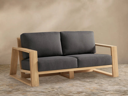 Canyon Outdoor Teak Sofa with natural teak wood frame and two dark gray cushions on a stone patio against a tan wall.