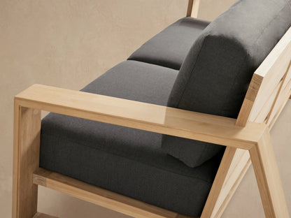 A two-seater Canyon Outdoor Teak Sofa with light teak wood frame and charcoal grey cushions on a stone patio.