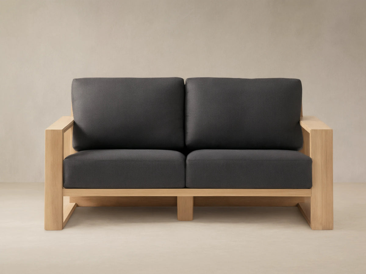 Canyon Outdoor Teak Sofa with natural teak wood frame and two dark gray cushions on a stone patio against a tan wall.