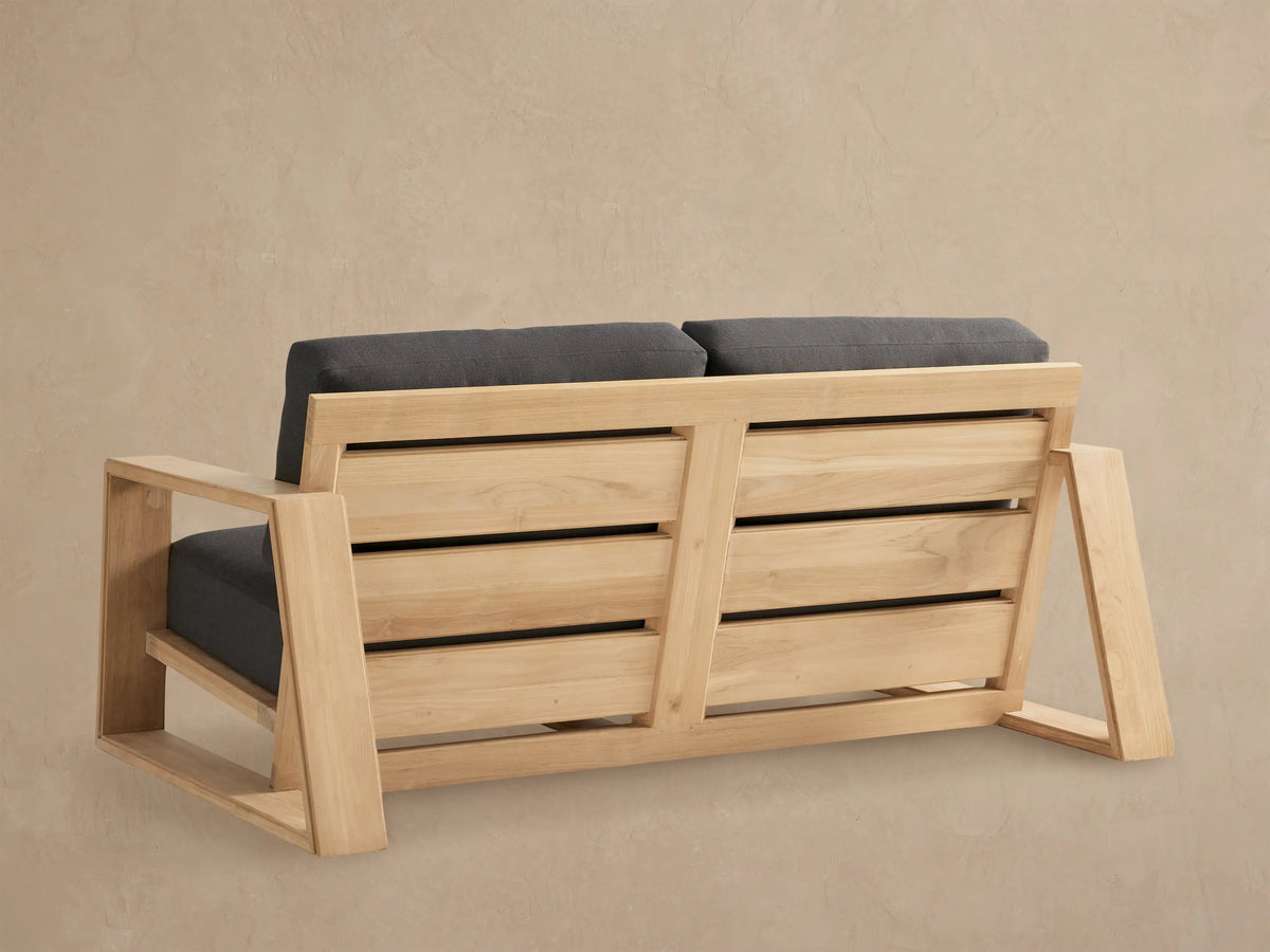 Angled view of Canyon Outdoor Teak Sofa. Light natural teak frame, square arms, and thick dark charcoal gray cushions on a beige background.