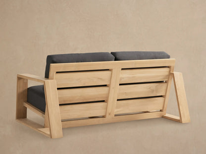 Angled view of Canyon Outdoor Teak Sofa. Light natural teak frame, square arms, and thick dark charcoal gray cushions on a beige background.