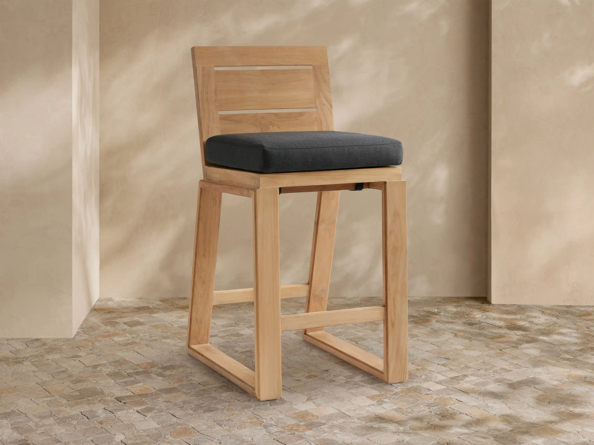 Canyon Outdoor Teak Barstool with a dark gray cushion, angled slightly, against a light tan wall & stone tile floor.