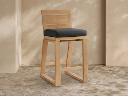 Canyon Outdoor Teak Barstool with a dark gray cushion, angled slightly, against a light tan wall & stone tile floor.