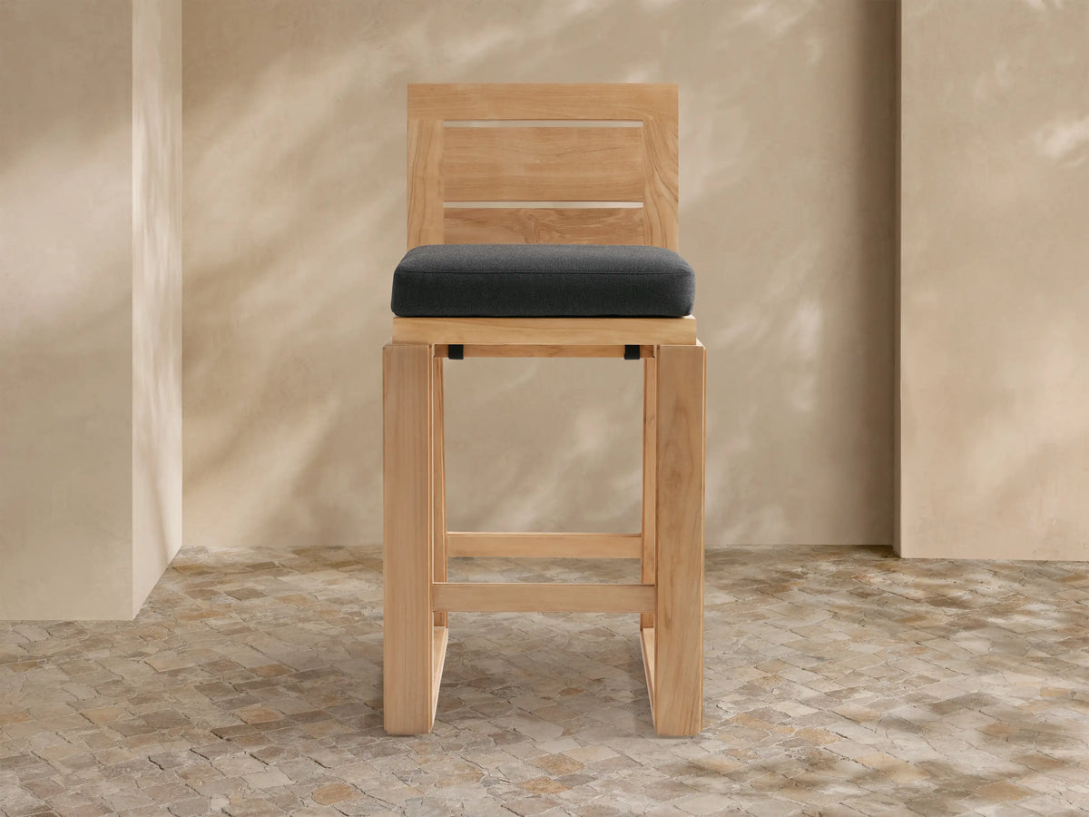 Canyon Outdoor Teak Barstool with a dark gray cushion, angled slightly, against a light tan wall & stone tile floor.