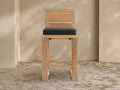 Canyon Outdoor Teak Barstool with a dark gray cushion, angled slightly, against a light tan wall & stone tile floor.