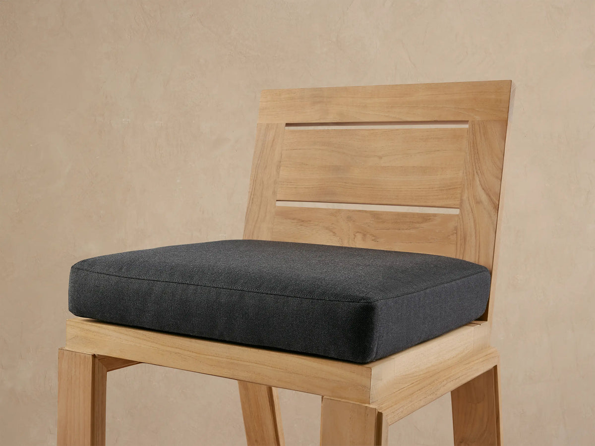 Front view of a natural teak Canyon Outdoor Barstool with a slatted back and dark gray cushion, on a textured patio.
