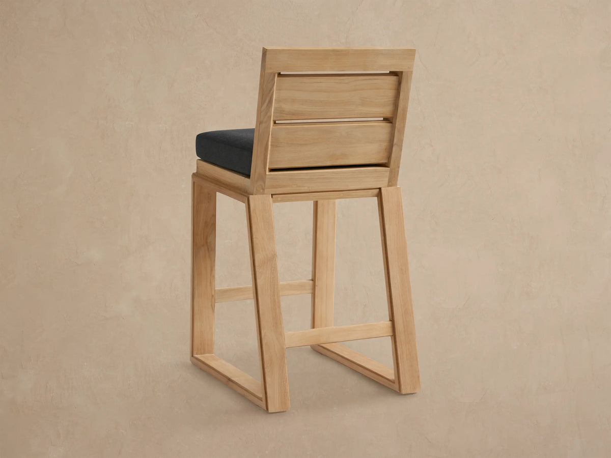 Canyon Outdoor Teak Barstool, light natural wood with slatted backrest, dark gray seat cushion. Angled view.