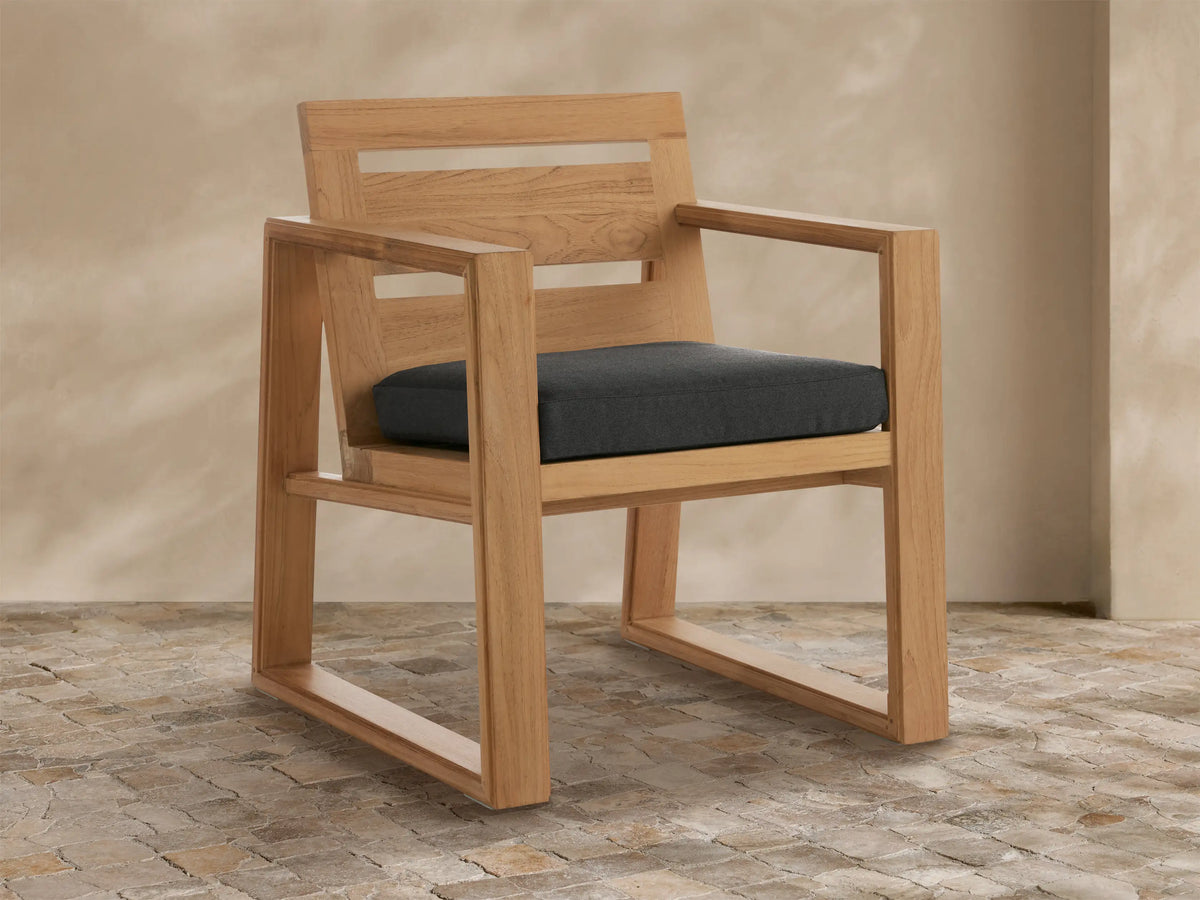 Canyon Outdoor Teak Dining Arm Chair, natural wood grain, dark grey cushion, with slatted backrest, on textured patio floor.