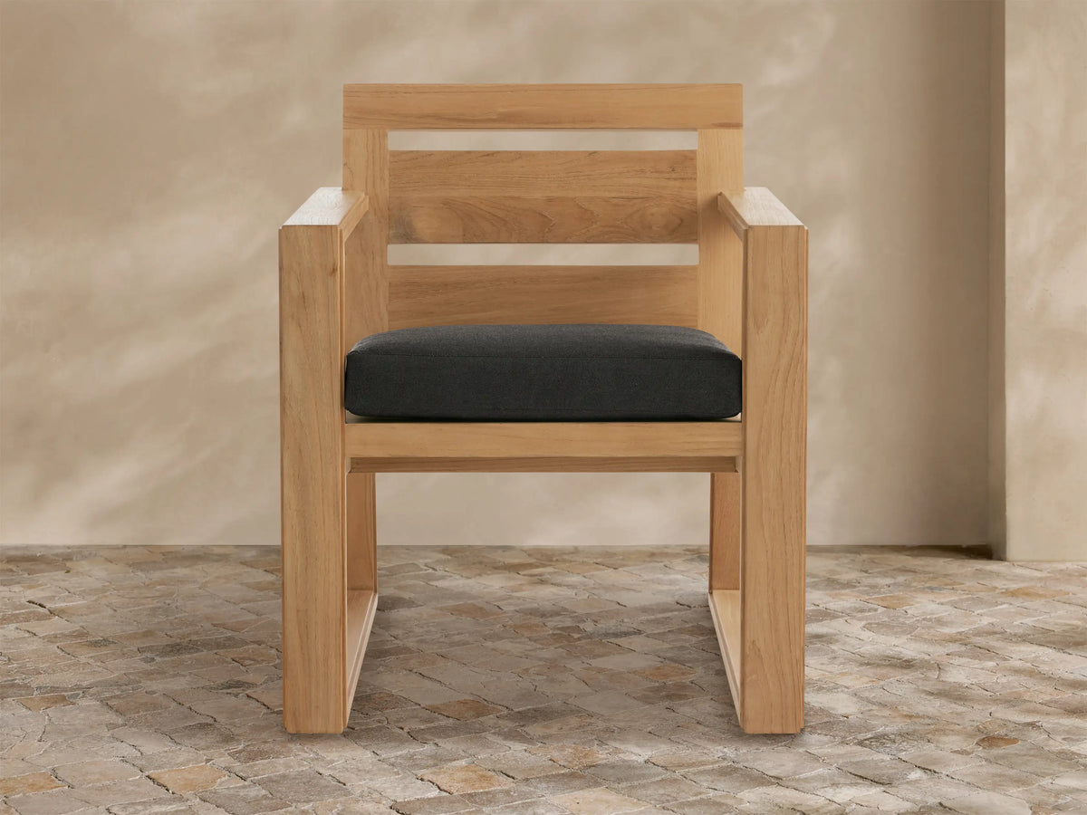 Canyon Outdoor Teak Dining Arm Chair, natural wood grain, dark grey cushion, with slatted backrest, on textured patio floor.