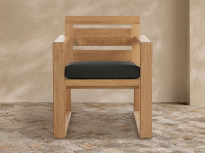 Canyon Outdoor Teak Dining Arm Chair, natural wood grain, dark grey cushion, with slatted backrest, on textured patio floor.
