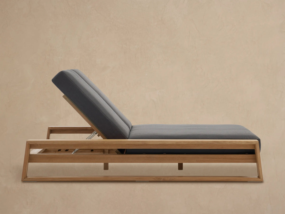 Canyon Outdoor Teak Double Chaise with a natural teak wood frame and dark gray cushions, backs reclined, on a patio.