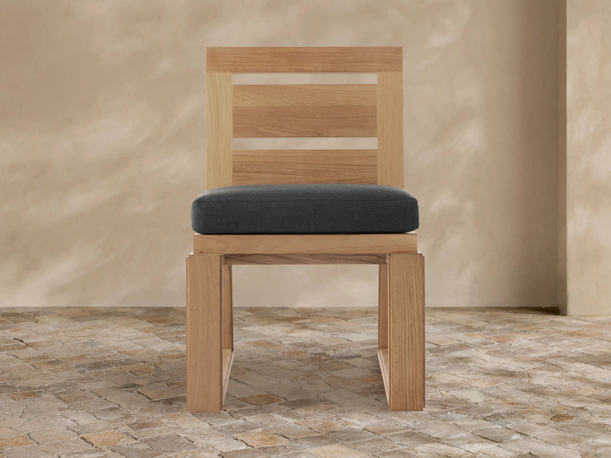 Canyon Outdoor Teak Dining Side Chair, natural light teak wood with a dark gray cushion, modern slatted back, on a rustic tiled floor.