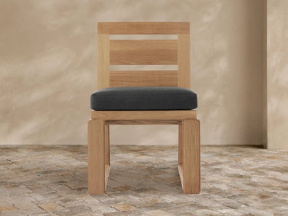 Canyon Outdoor Teak Dining Side Chair, natural light teak wood with a dark gray cushion, modern slatted back, on a rustic tiled floor.