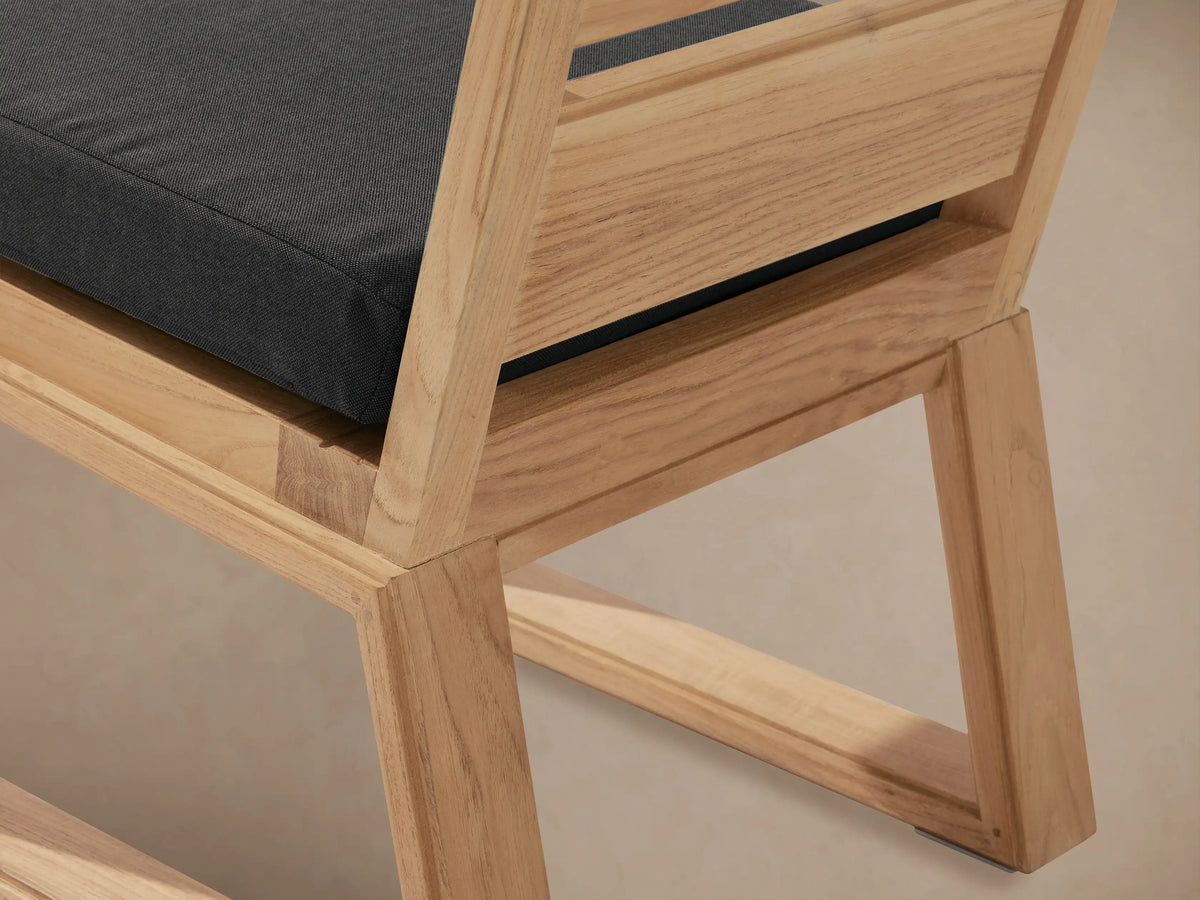 Canyon Outdoor Teak Dining Side Chair: front view of light natural teak wood, slatted back, thick dark grey cushion on seat, on tiled floor.