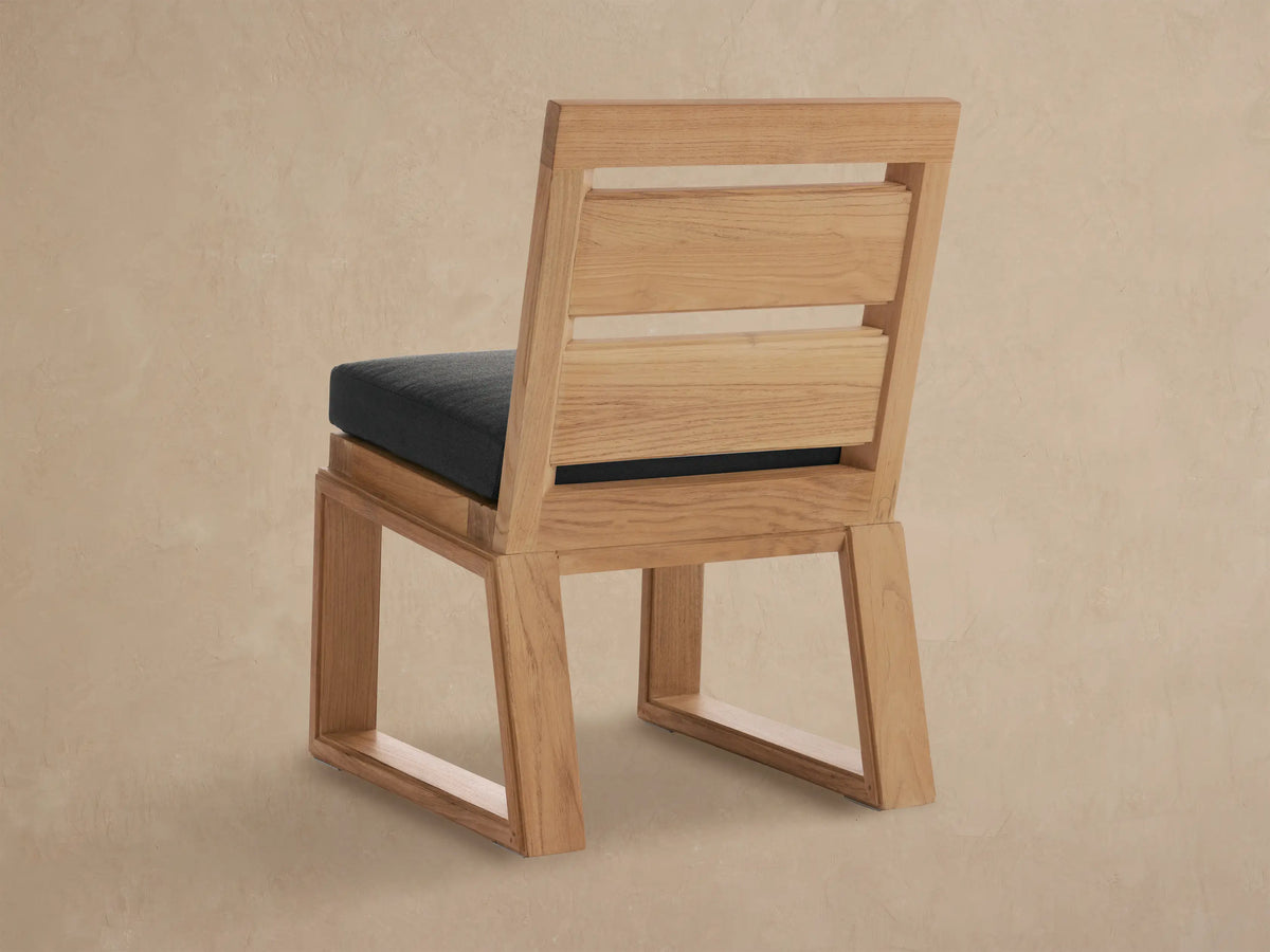 Close-up of Canyon Outdoor Teak Dining Side Chair, showing natural light brown teak wood frame and dark gray seat cushion at an angle.