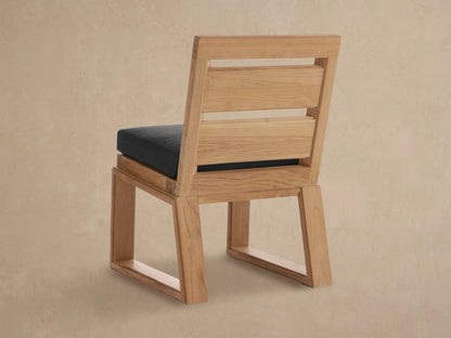 Close-up of Canyon Outdoor Teak Dining Side Chair, showing natural light brown teak wood frame and dark gray seat cushion at an angle.